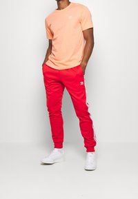 Muž oblečený v korálovém tričku Adidas a červených teplácích Adidas s bílými pruhy, stojící se zhandy v kapsách proti bílému pozadí.