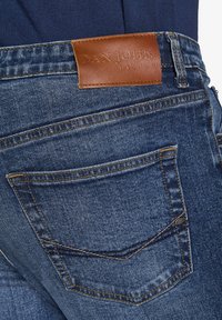 I jeans in denim blu scuro presentano un'etichetta in pelle marrone sulla vita con la scritta "DAN JOHN" impresso. I dettagli delle cuciture enfatizzano il design.