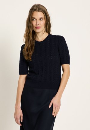 Frau mit langen, welligen Haaren, die einen marineblauen kurzärmligen Pullover mit Zopfmuster und einen passenden marineblauen Rock trägt, vor einem schlichten Hintergrund stehend.