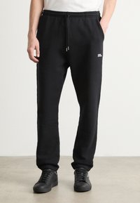 Svarte joggebukser laget av mykt stoff, med elastisk midje med snøring, strømper ved ankelen, og en liten hvit logo på siden.