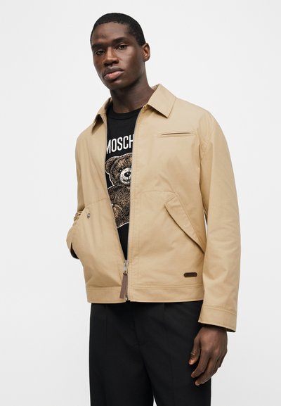 Άνδρας με μπεζ μπουφάν με φερμουάρ πάνω από μαύρο μπλουζάκι με γραφικό αρκούδας και τη λέξη "MOSCHINO", συνδυασμένο με μαύρο παντελόνι.