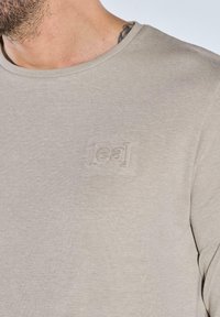 Helles graues T-Shirt aus weichem Stoff mit Rundhalsausschnitt und einem geprägten [eal] Logo auf der Brust. Kurze Ärmel und glatte Textur.