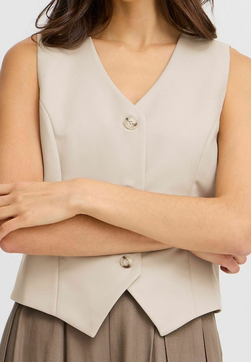 Gilet sans manches beige avec un col en V et deux boutons à l'avant. Coupe cintrée montrée avec les bras croisés, affichant une texture lisse.