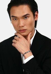 Silver chain bracelet featuring rectangular links, worn on a wrist, with polished surfaces and decorative accents, against a black suit jacket.