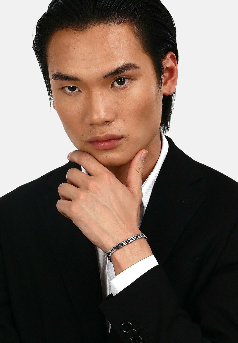 Silver chain bracelet featuring rectangular links, worn on a wrist, with polished surfaces and decorative accents, against a black suit jacket.