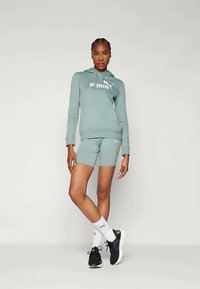 Puma kapuutsiga sviiter ja lühikesed püksid tuhmroheline kangas, esiküljel logo, tasku ja sportlik disain. Mustad tossud ja valged sokid.