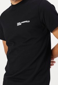 Černé bavlněné tričko s kulatým výstřihem a krátkými rukávy. Bílý nápis "KARL LAGERFELD JEANS" na levé straně hrudi. Hladký materiál, standardní střih.