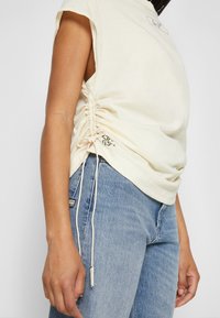 Krémový top na krátký rukáv s bočními řasením a šňůrkami. Nosený s světle modrými džínami. Obsahuje malý grafický detail na lemu.