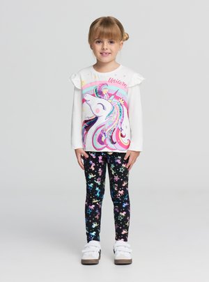 Ragazza giovane in piedi che sorride, indossa una maglia bianca a maniche lunghe con unicorno e leggings neri con motivi colorati di unicorni e stelle, scarpe da ginnastica bianche.