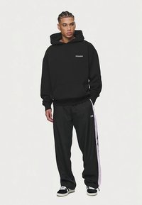 Melns kapuces džemperis ar priekšpusi kabatu un baltu "PECADOR" logo, kopā ar melnām sporta biksēm ar baltu sānu joslu.