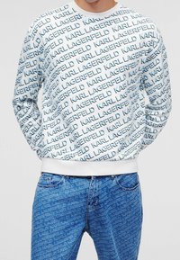 Felpa bianca con un motivo testuale ripetuto in blu "KARL LAGERFELD", con polsini e orlo a costine. Abbinata a jeans blu con un design simile.