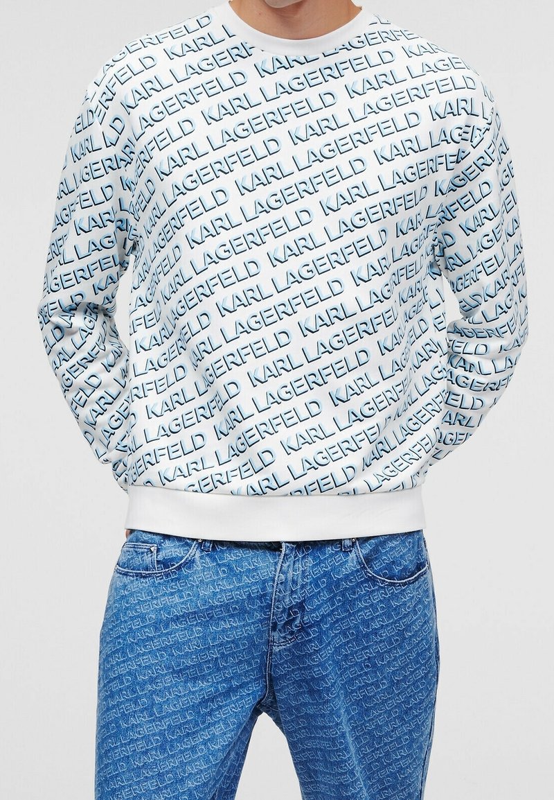 Felpa bianca con un motivo testuale ripetuto in blu "KARL LAGERFELD", con polsini e orlo a costine. Abbinata a jeans blu con un design simile.