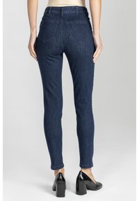 Kobieta ubrana w ciemnoniebieskie slim fit dżinsy oraz czarne buty na niskim obcasie, stojąca z rękami swobodnie u boku, zwrócona plecami na jednolitym tle.