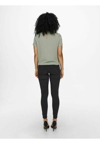 Top verde oliva a maniche corte con dettaglio a chiave sul retro, abbinato a jeans skinny neri e scarpe con tacco alto nere. Design semplice.