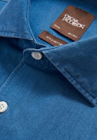 Chemise en denim bleu moyen, avec un col boutonné, des boutons blancs et une étiquette portant la marque "Oscar Jacobson". Texture lisse.