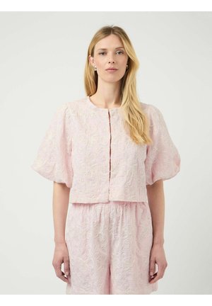 Kvinde med langt blond hår iført en lyserød blondebluse med pufærmer og matchende blonde-shorts mod en ensfarvet baggrund.