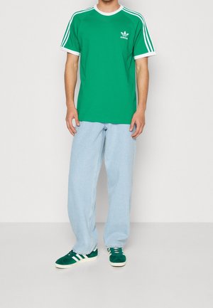 Mand iført grøn Adidas t-shirt med hvide striber og logo, lyseblå jeans med vide ben og grønne Adidas-sneakers med hvide striber.