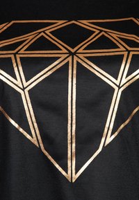 Černé tričko z látky s velkým geometrickým diamantovým designem v metalické zlaté barvě, který vykazuje ostré linie a hladkou texturu.