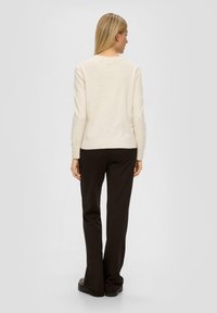 Hellbeige Strickpullover mit langen Ärmeln und Knopfdetails an den Bündchen, kombiniert mit dunkelbraunen Schlaghosen und schwarzen flachen Schuhen.