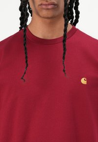 Burgundy cotton t-shirt featuring a round neckline and short sleeves, with a small gold logo on the left chest. Plain texture.