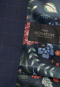 Marineblaues Textil mit einem Karomuster, kombiniert mit einem blumigen Satinfutter in Blau, Grün und Rot. Das Etikett lautet "next SIGNATURE PREMIUM TAILORING 40R."