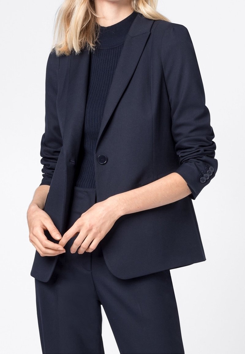 Námořnický modrý blazer s klopami, zapínání na jeden knoflík a přiléhavý střih. Měkká, texturovaná látka; ohrnuté rukávy s akcenty na knoflíky na manžetách.