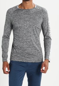 Man wearing a heather gray long-sleeve shirt with white text on one sleeve and blue pants, standing against a plain background.