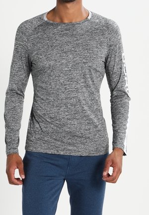 Mann trägt ein meliertes graues Langarmshirt mit weißem Text auf einem Ärmel und blaue Hosen und steht vor einem schlichten Hintergrund.