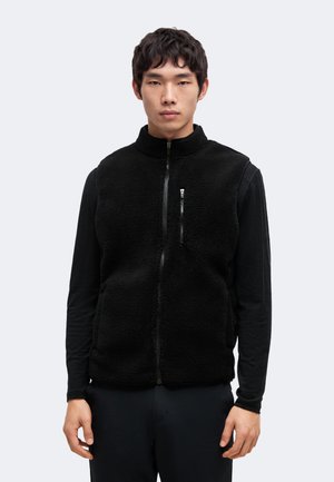 Un homme portant un gilet polaire noir zippé par-dessus une chemise à manches longues se tient devant un fond clair et uni.