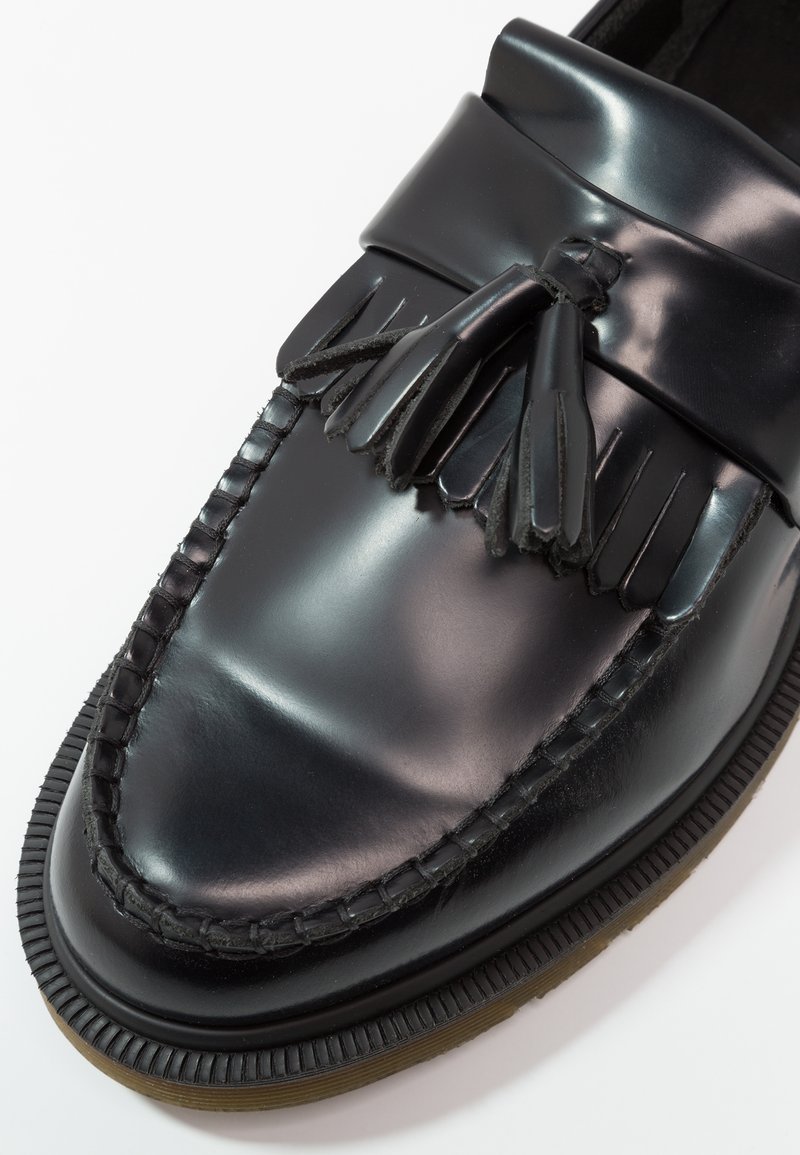 Mocassino in pelle nera con finitura liscia, caratterizzato da una tomaia intrecciata, dettagli con frange e una suola in gomma spessa. Gli accenti includono cuciture decorative.