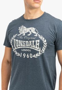 Camiseta de color azul marino hecha de algodón, con un gráfico de un león blanco y el texto "The Original Lonsdale London 1960", con un diseño de cuello redondo.