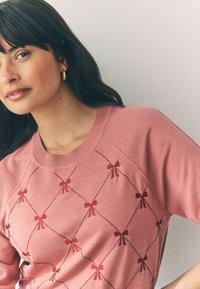 Rosa Pullover mit einem strukturierten Design, das rote gestickte Schleifen in einem Rautenmuster aufweist. Weicher Stoff und klassischer Rundhalsausschnitt mit geripptem Saum.