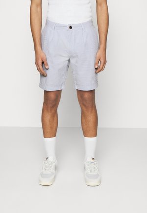 Homme portant un short à carreaux bleu clair et blanc, un débardeur blanc, des chaussettes blanches et des baskets blanches, debout sur un fond blanc.