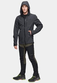 Czarna kurtka softshell z kapturem, z zamykanymi kieszeniami; zielone i czarne spodnie z wzmocnionymi elementami; czarne i zielone buty trekkingowe na sznurówki.