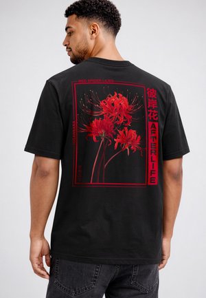 Mann, der mit dem Rücken zum Betrachter steht, trägt ein schwarzes T-Shirt mit roten Spinnenlilienblumen und rotem Schriftzug „Afterlife“ sowie japanischen Zeichen auf dem Rücken.