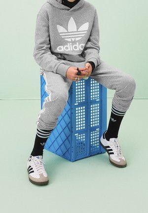 Sudadera gris con gran logo blanco de Adidas, pantalones de chándal a juego, calcetines negros con rayas y zapatillas blancas con detalles marrones, sentado en una caja azul.