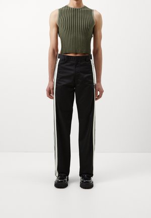 Schwarz eng anliegende Hose mit weißen Seitenstreifen, kombiniert mit einem ärmellosen, olivgrünen gerippten Croptop und schwarzen klobigen Schuhen.