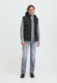 Personne souriante portant un gilet rembourré noir sur un pull gris, un jean clair et des bottes noires, sur un fond clair et uni.