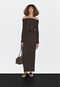 Frau mit lockigem Haar, die ein braunes, schulterfreies Strickkleid, eine schwarze Anhängerhalskette trägt, eine braune Handtasche hält und schwarze Sandalen trägt.