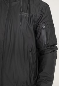 Czarna kurtka bomberka ma gładkie, matowe wykończenie, boczne kieszenie oraz kieszeń na rękawie zapinaną na zamek. Zawiera regulowany kaptur ze sznurkiem.