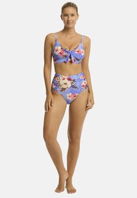 Model poseert in een bloemenbikini met strikdetail. De achtergrond is helder wit, wat de levendige kleuren van het zwemkleding benadrukt.