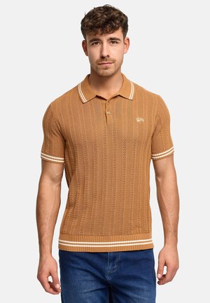 Młody mężczyzna stoi, ubrany w brązową teksturowaną koszulkę polo z krótkim rękawem, z białymi paskami na rękawach i na dole oraz w niebieskie dżinsy na jasnym tle.