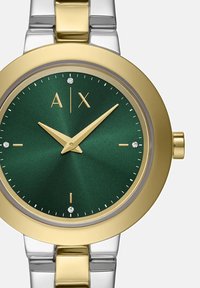 Kuld- ja hõbetooniline kell rohelise näo, kuldsete kätega, mis on kell 10:10 asendis, teemantist tunni markeerijad ja "A|X" logo kell 12 positsioonil.