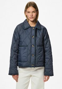 Navy spalvoto pamušalo švarkas su dideliu apykakle, dviem priekinėmis kišenėmis ir dideliais mygtukais. Turėdamas dėžinę formą ir sklandžią, blizgią tekstūrą.