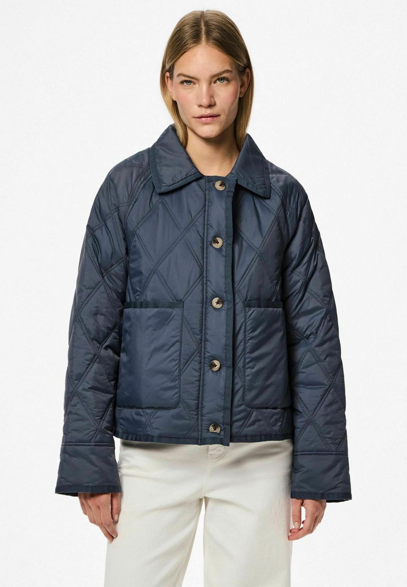 Navy spalvoto pamušalo švarkas su dideliu apykakle, dviem priekinėmis kišenėmis ir dideliais mygtukais. Turėdamas dėžinę formą ir sklandžią, blizgią tekstūrą.