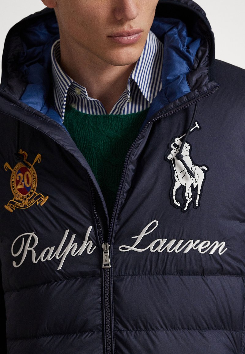 Male wearing navy Ralph Lauren puffer jacket with embroidered logo, crest patch, green cable-knit sweater, and blue striped collared shirt.