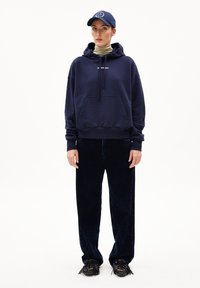 Felpa navy con tasca frontale e logo, abbinata a pantaloni in corduroy a gamba larga di colore blu scuro e scarpe nero con dettagli frange.