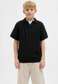 Polo noir à manches courtes avec un col côtelé, présentant des rayures verticales texturées et une coupe ample. Porté par-dessus une chemise blanche.