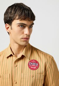 Beżowa koszula z guzikami w zielone paski, z okrągłą czerwoną naszywką z napisem "CASEY JONES" oraz "Wrangler", posiadająca dwie przednie kieszenie.