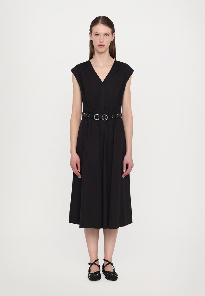 Vestido preto até ao joelho com decote em V, mangas curtas, corpete pregueado e um cinto largo com detalhes metálicos circulares. Sapatos pretos com tiras.